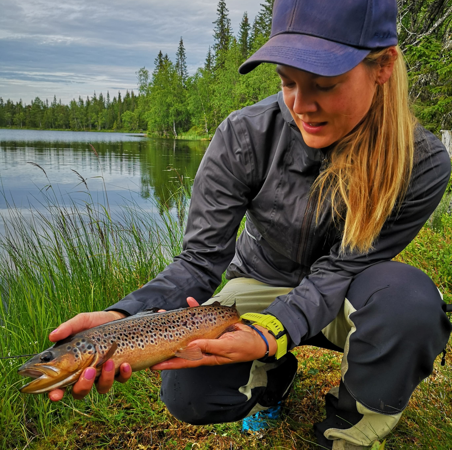 Bilde av Wenche Christenssen i Fiskejenter i Marka som sitter på huk ved bredden av et vann og holder en brun ørret i hendene. Hun smiler og ser ned på fisken hun har fanget. Hun har blå caps, lang blond hestehale og friluftsklær på seg. 